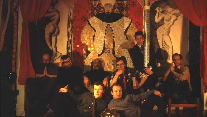 Group portrait of nine people posing before a large ornate mural with theatrical red curtains and a "1968" sign in front.