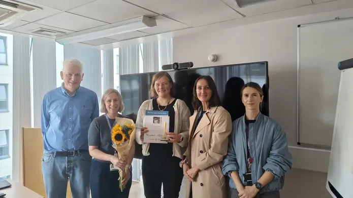 Esther Sammler receiving her award and a presentation of flowers from well-wishers, standing in a classroom environment