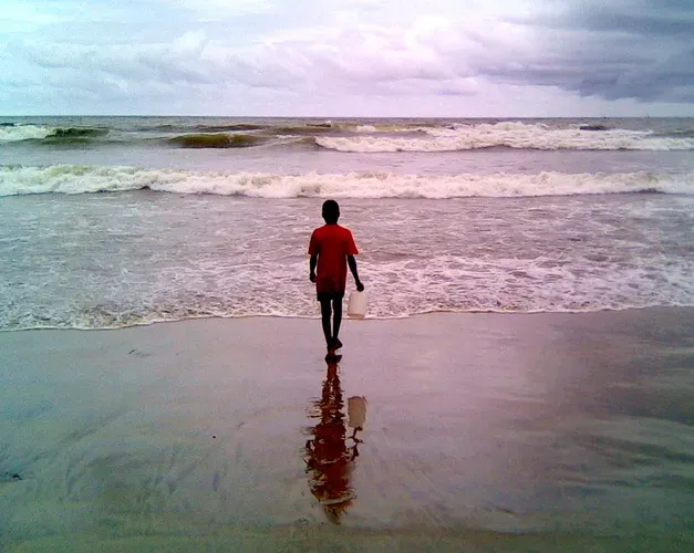 A boy is walking toward the shoreline on a beach, with a white bucket in his hand