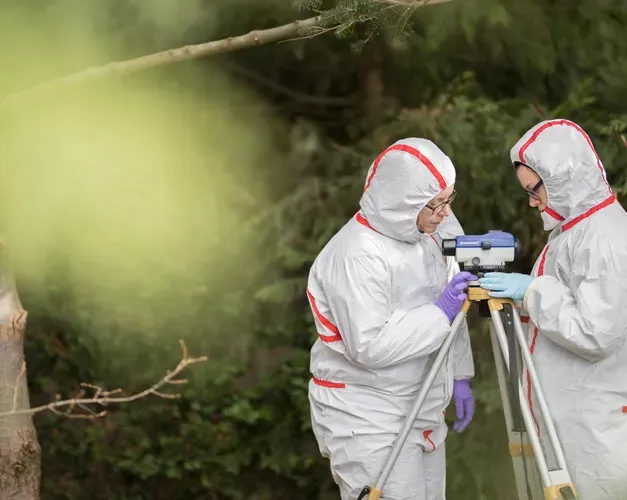 two people in full body clean suits look at a cameralike device on a tripod in the woods