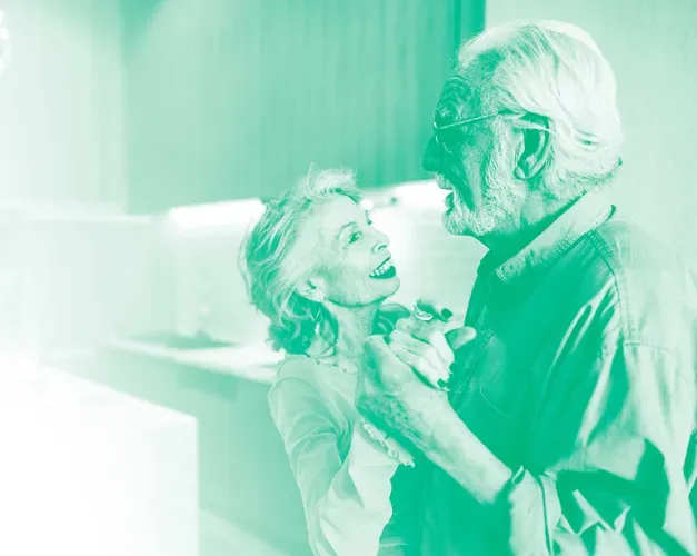 An elderly couple dance in their kitchen with a green colour wash over the photo