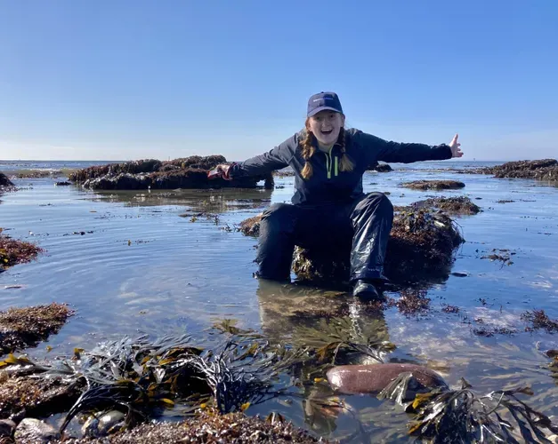 a young woman in her 20's surrounded by rockpools of shimmering silver and blue sea water on a clear, blue skied day