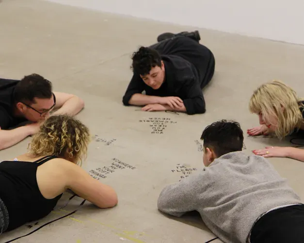People lying face down on the floor in a circle looking at writing on the floor