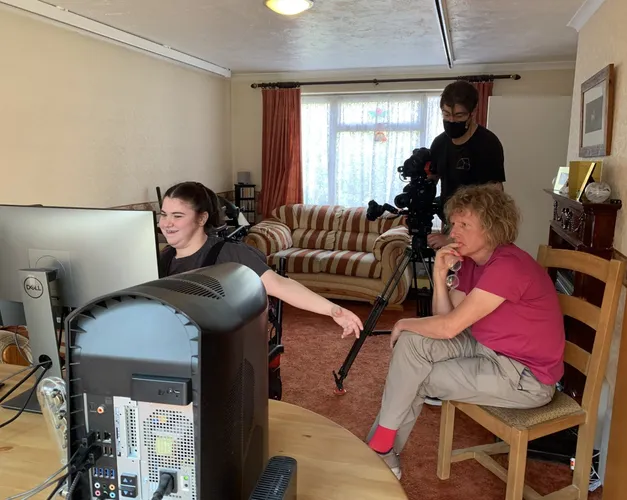 a middle aged man sat on wooden chair, looking with interest at a young, late-teened girl sat in a wheelchair as she works on a computer