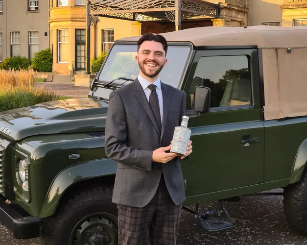 Lewis Kenned holds a bottle of his non-alcoholic gin with a Land Rover and traditional Scottish country hotel behind him