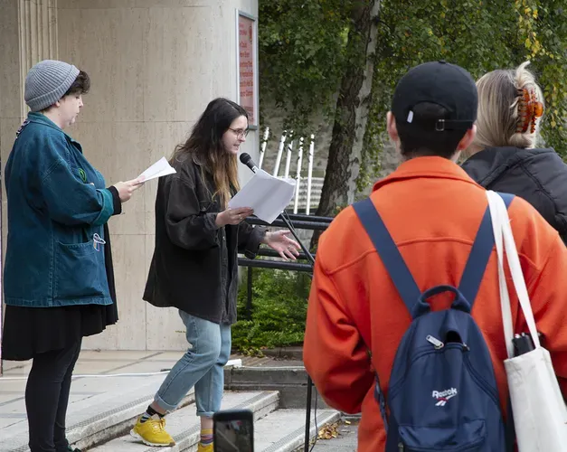 Audiences with Three white performers reading from scripts into a single microphone on the steps of the gallery underneath a decimal clock
