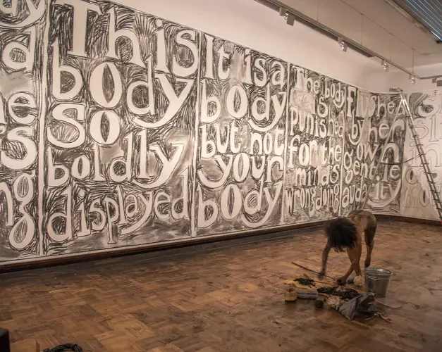 Large words are written in charcoal on a gallery wall. An artist is in the space without clothes working on the drawn lettering. The charcoal soot is everywhere in the space. 