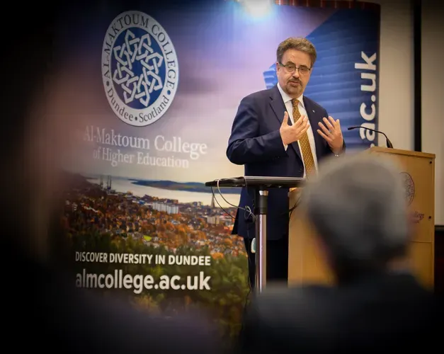middle aged man standing on stage behind podium, wearing dark navy suit and speaking to a crowd of people who can be seen in foreground