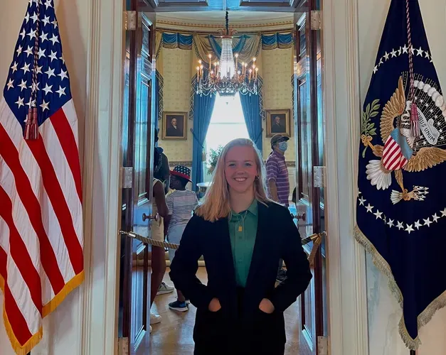 Young girl in her early 20's wearing a black suit jacket stands posing in front of an open door to the Oval Office, which is the office of the President of the USA. On her left a large American flag hangs down and on her right is the President Seal flag