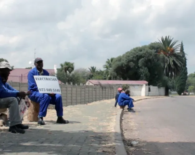 Black workers in blue overalls waiting in a road side with a sign reading electrician and a phone number