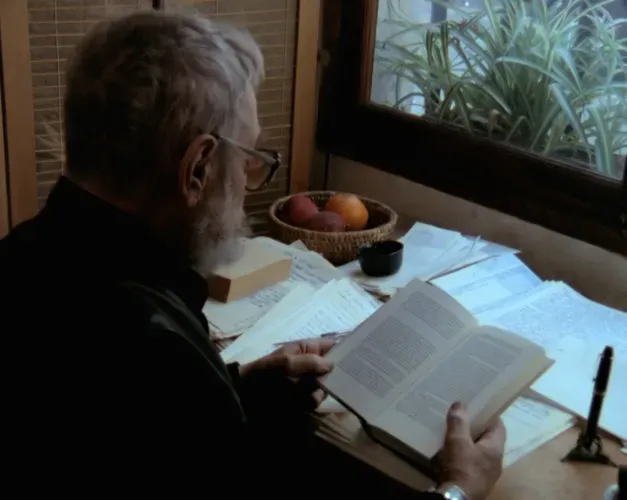 Film still. An elderly man with grey hair sits reading a book at a writing desk. 