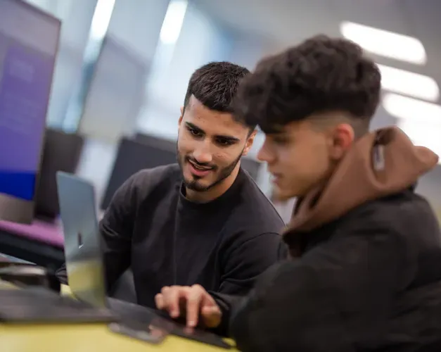 two students work on a laptop together, other computers are in the background along with large monitors