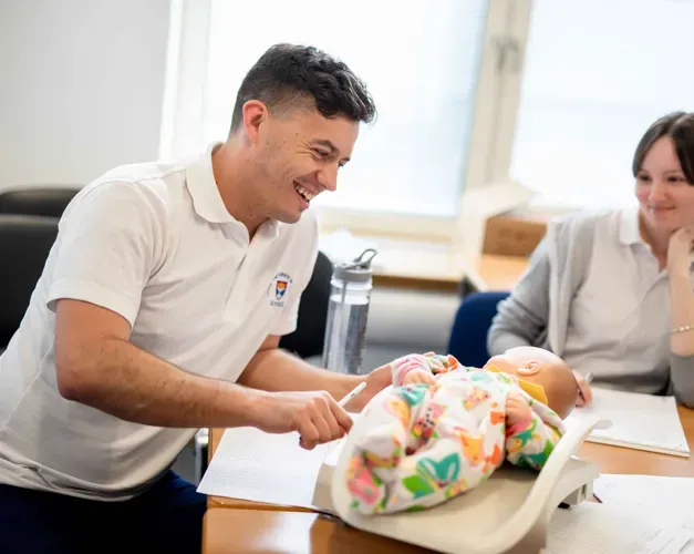 A male and female, both in Dundee University branded shirts are sitting at a table with a baby doll between them.