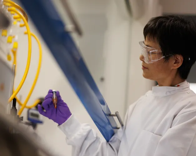 A person wearing purple gloves and goggles in a lab looking at a test tube