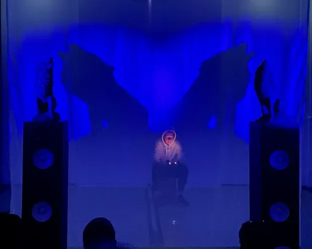 Heather Phillipson reading lit by the orange glow of her phone. She is bathed in blue light and flanked by two howling wolf sculptures and their looming shadows.