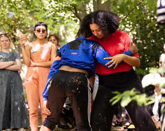 Two performers leaning on each other in a shady wooded area surrounded by audiences