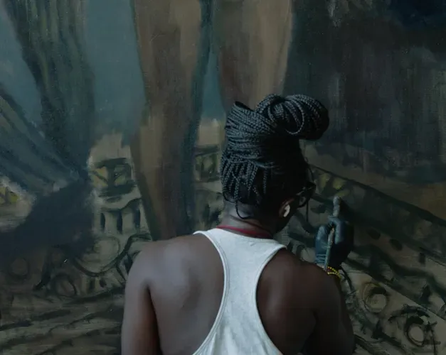 A black woman painting on a large canvas showing the legs of two people on a rug. The painter has a white vest and her braids are bunched in a bun.