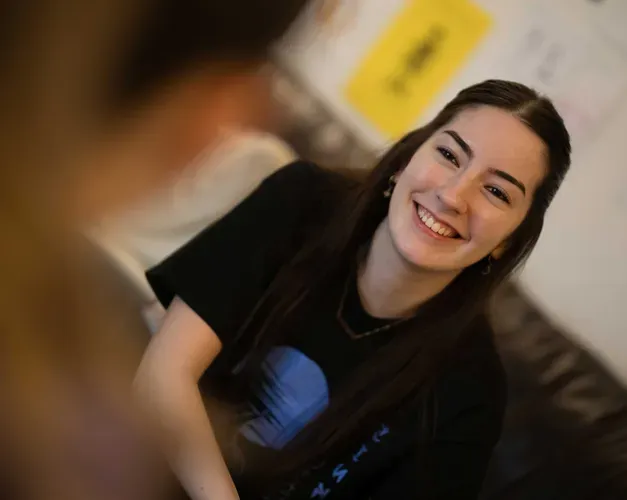 A girl with long dark hair smiles towards a person who is blurred in the foreground.