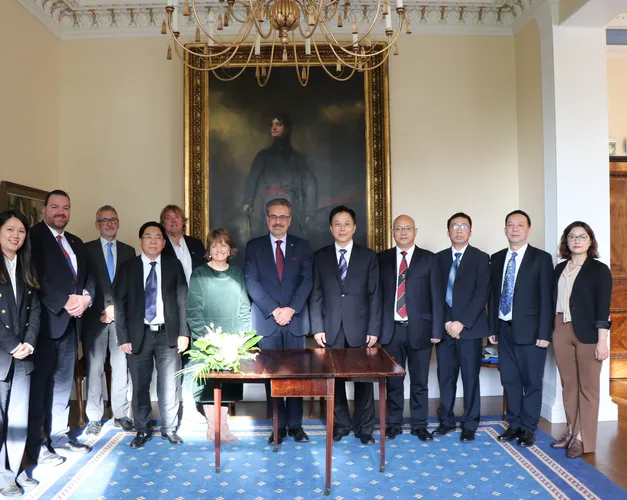 Group of men and women in a stately room in front of a large painting with a brown table with flowers on it in front of them.
