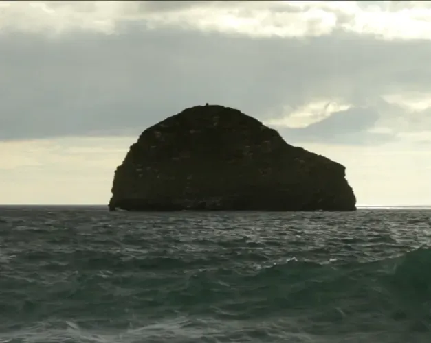 Film still. A rock in the ocean sits under a cloudy sky surrounded by a rough sea
