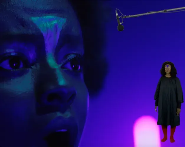 A close up of a black woman's face with a triangle painted on her forhead. Another woman with a triangle on her forehead is superimposed standing full height next to the looming head. A mic boom cuts across the image, the set is purple.