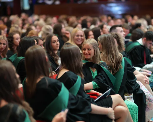 A group of students in black graduation robes with green hoods sitting in a theatre smiling at eachother