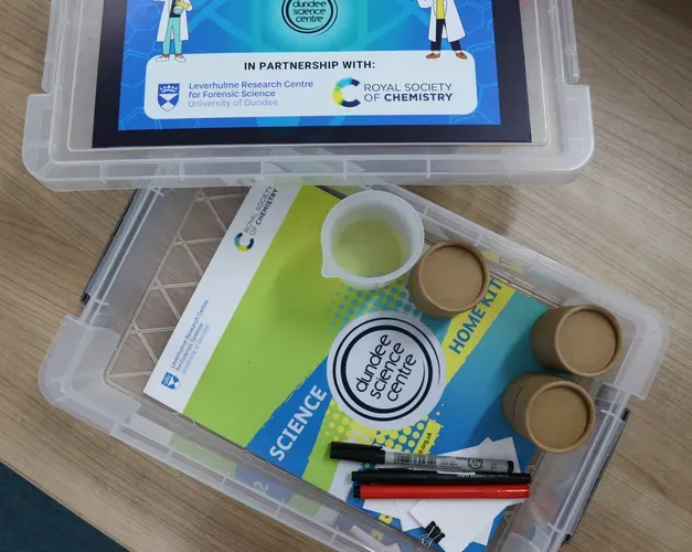 Storage box and lid on a wooden table with science equipment and a green and blue leaflet inside.