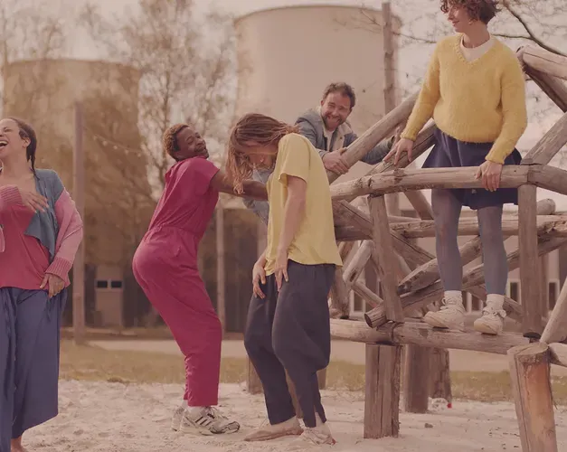 Film still. Six dancers in brighly coloured clothing hang out in a public park