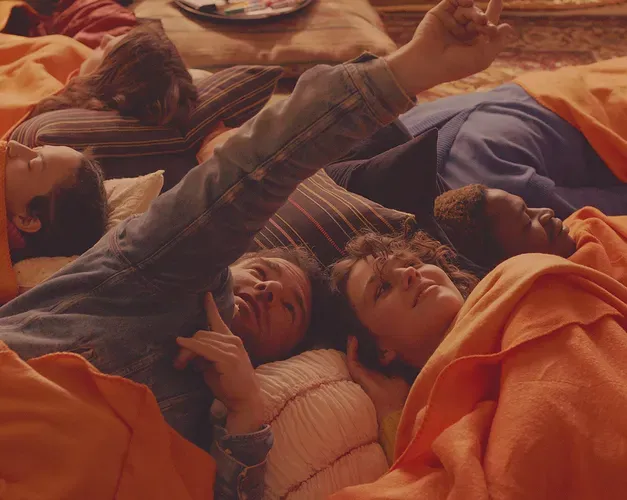 Film still. A group lie down wrapped in orange blankets, their heads almost touch forming a cirle