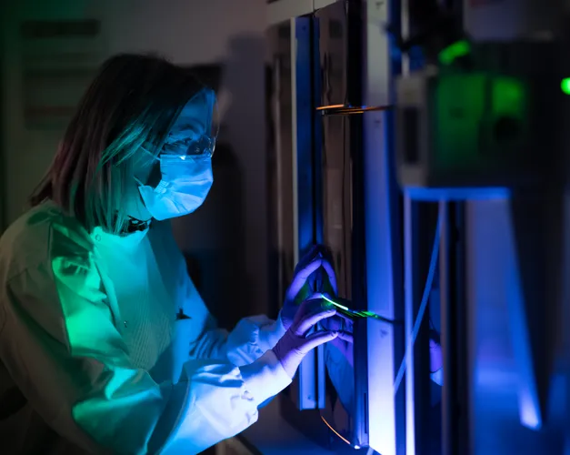 a women in a lab coat and mask is lit with blue and green lab as she does work in the lab