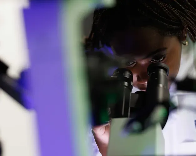 A woman in a lab coat looks through a microscope. A blue-green line cuts across the image.