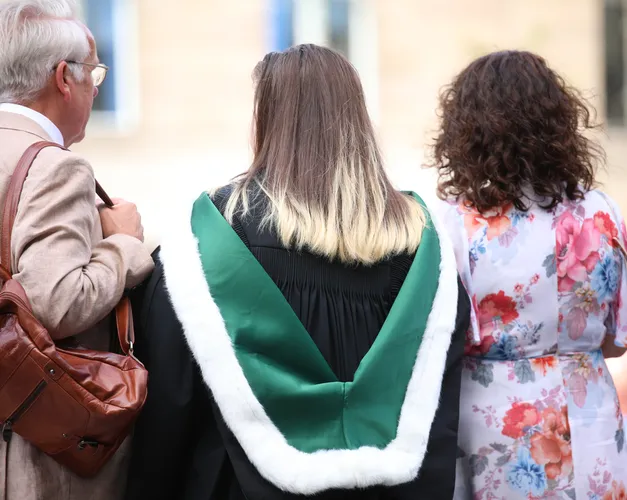 Three people standing with their back to the camera. Young female in centre wearing a graduation gown and green hood lined with white fur. Older male on left holding brown bag over his shoulder and older woman on first hand side wearing floral dress.