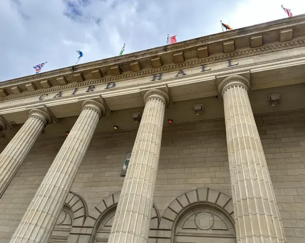Roof area of old building with tall pillars and flags out of the top, and a cloudy blue sky above