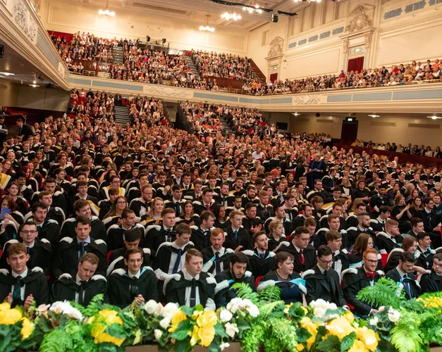 Hundreds of students sitting together in a large hall for their graduation ceremony. There is a upper level for their families.  