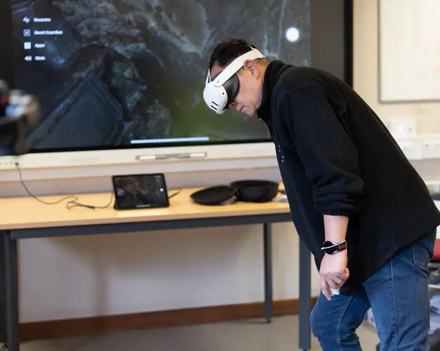 Man wearing virtual reality headset bending to side with screen behind him in classroom setting