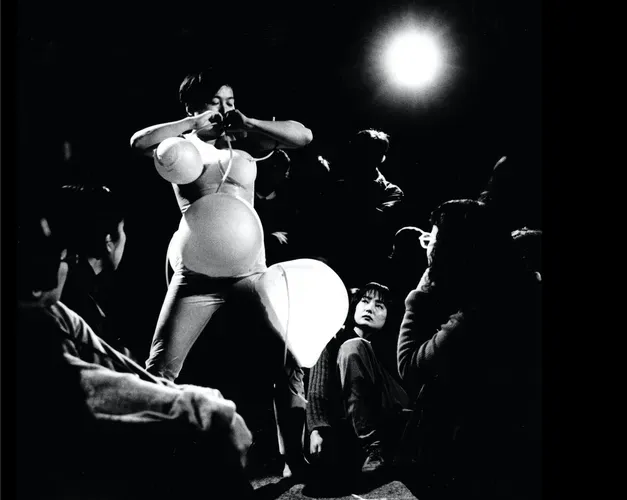 Tari Ito performance view black and white. Artist blowing inflatables around her body with tubes connected to her mouth creating enlarged spheres on her stomach, chest
