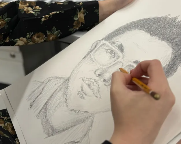 Close up of an unseen individual's hands sketching a face of a man with a pencil