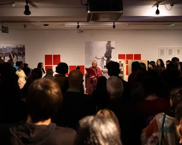 A group of people watching a woman speak in front of an art gallery exhibition