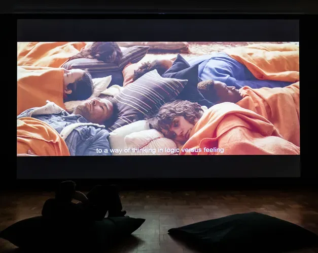 Person in a darkened room watching a large projection on a bean bag. The screen is the size of a double decker bus and shows a circle of people lying together on orange bean bags