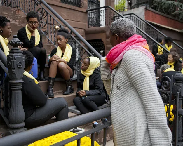 A group of women sit on the steps of a New York flat talking. A passerby stands and listens. The group talking wear yellow scarves