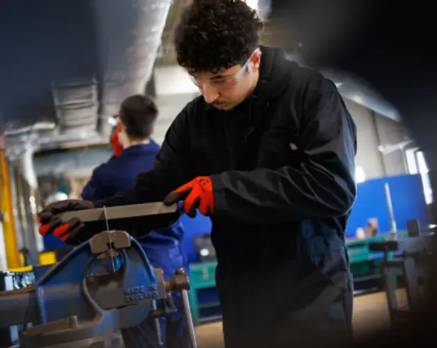 A student in a boiler suit and gloves works a piece of metal being held in a vice