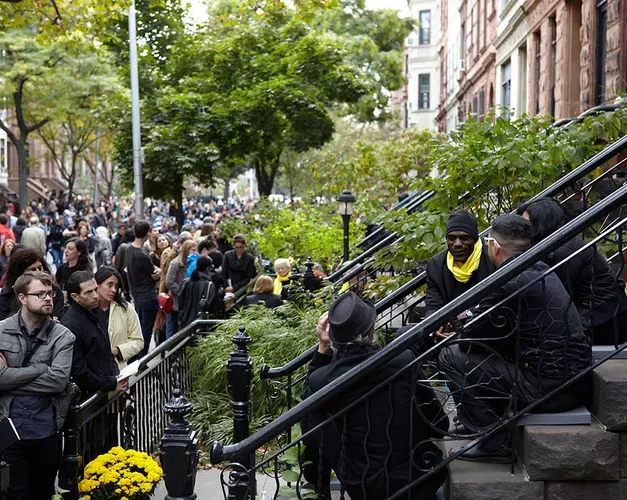 large crowd gather on a new york street, on a stoop are a group of women sitting and talking wearing yellow scarfs