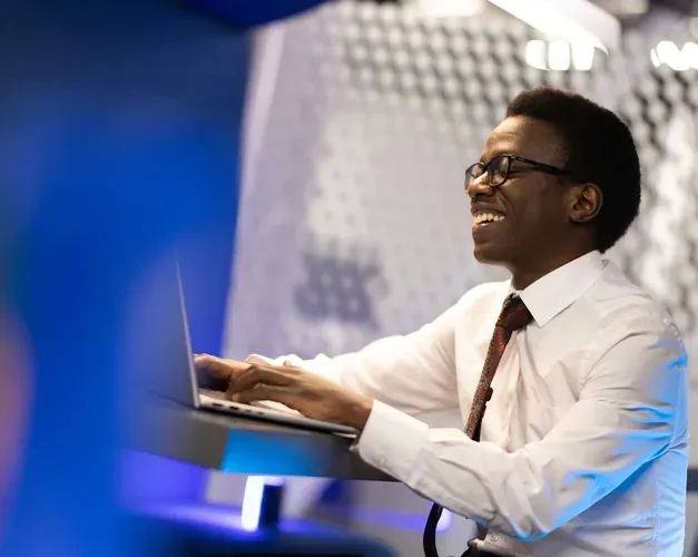 A man in a white shirt and black tie sits at a laptop, smiling.