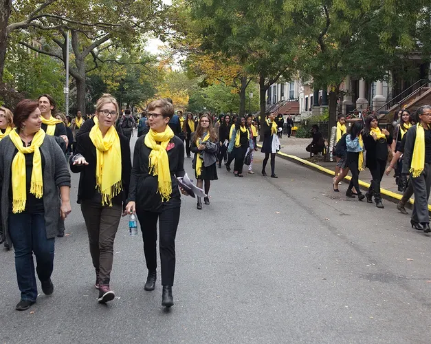 Many women walk down a Brooklyn street wearing yellow scarves, two women at the front are walking and talk