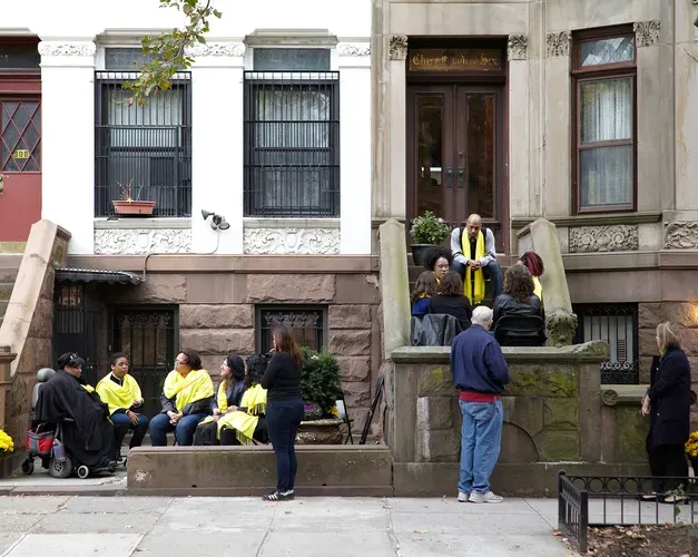 Performance photograph. Groups sit on the front steps of a new york street in discussion, performers wear yellow scarves