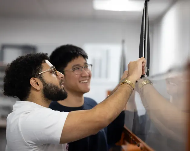 Two people stand together in front of a large piece of glass, one of them holds a device against it.