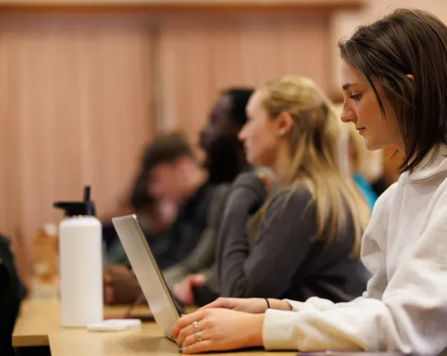 Side view of a young woman sat at a desk with a laptop open looking a the screen. Others sat next to her also look to be working