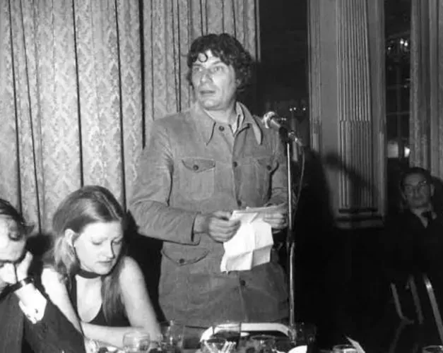 Black and white photography of John Berger, a white middle-aged-man, standing at an awards ceremony, speaking to the audience seated around tables. He holds a piece of paper he is reading from.