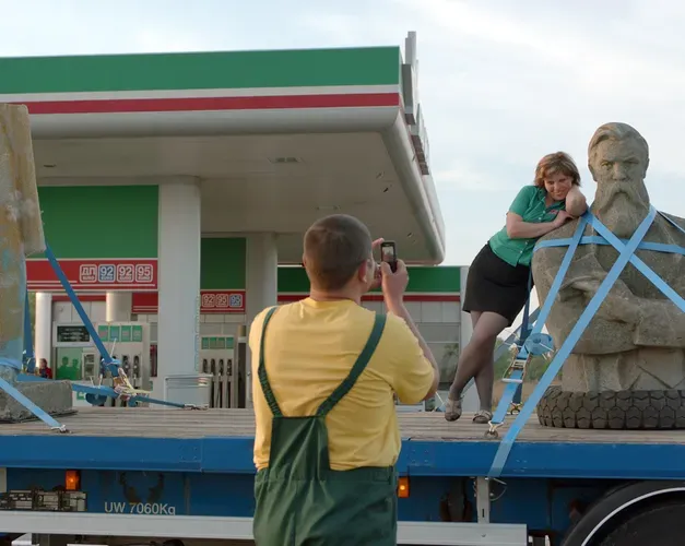 Film still. A sculpture of Frederick Engels is strapped to the back of an open topped truck. It is stopped at a petrol station. A woman leans on the status' shoulders while a man takes a photo of her on the truck leaning on Engels