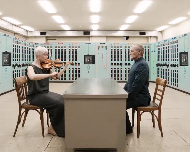Film still. Two protagonists sit facing each other across a table. One is playing a violin and the other is dressed as a diplomat. They are in an engine control room surrounded by switches and buttons